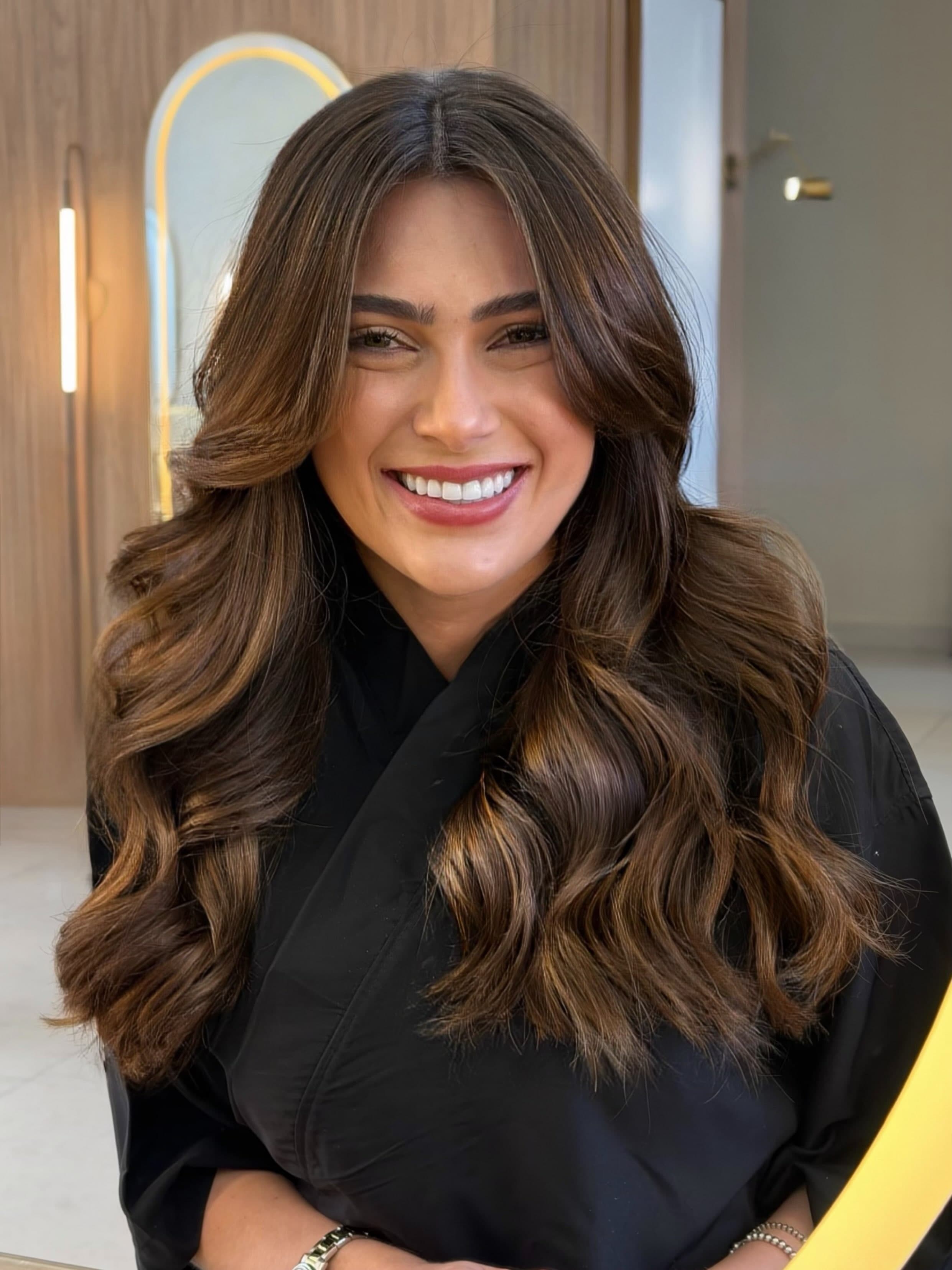 Cliente sorrindo com o cabelo castanho caramelo em ondas, diante de um espelho oval